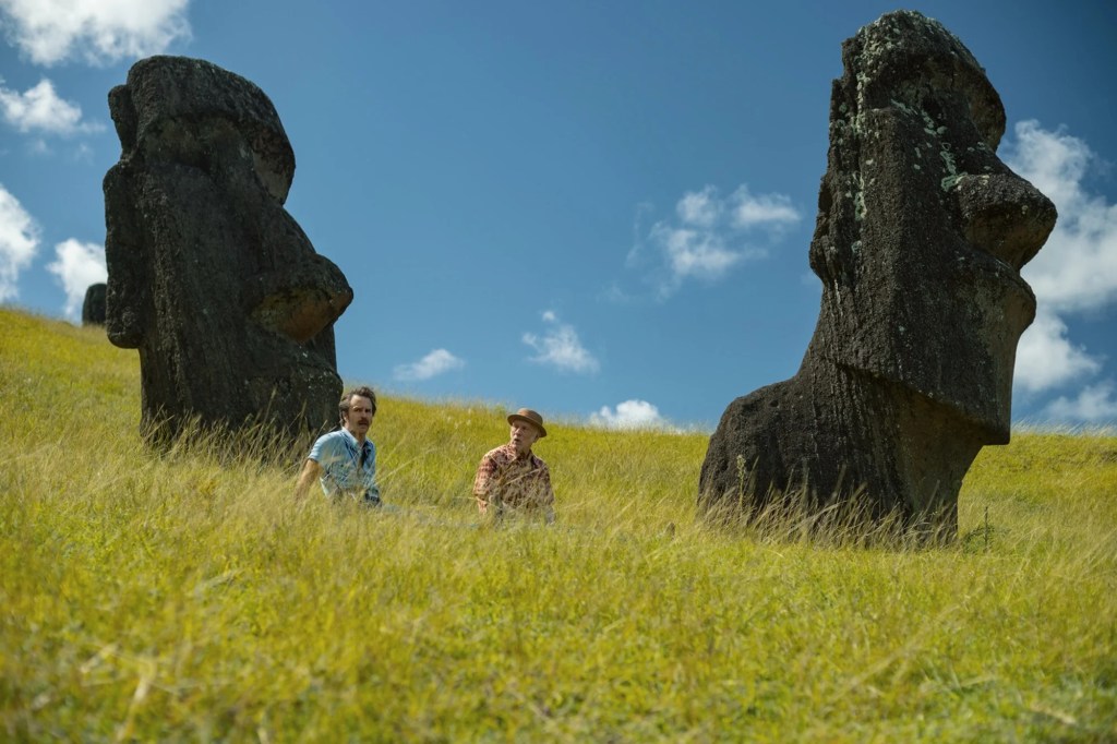 Chris and Lee sitting in tall grass on a hillside between two giant moai statues on Rapa Nui