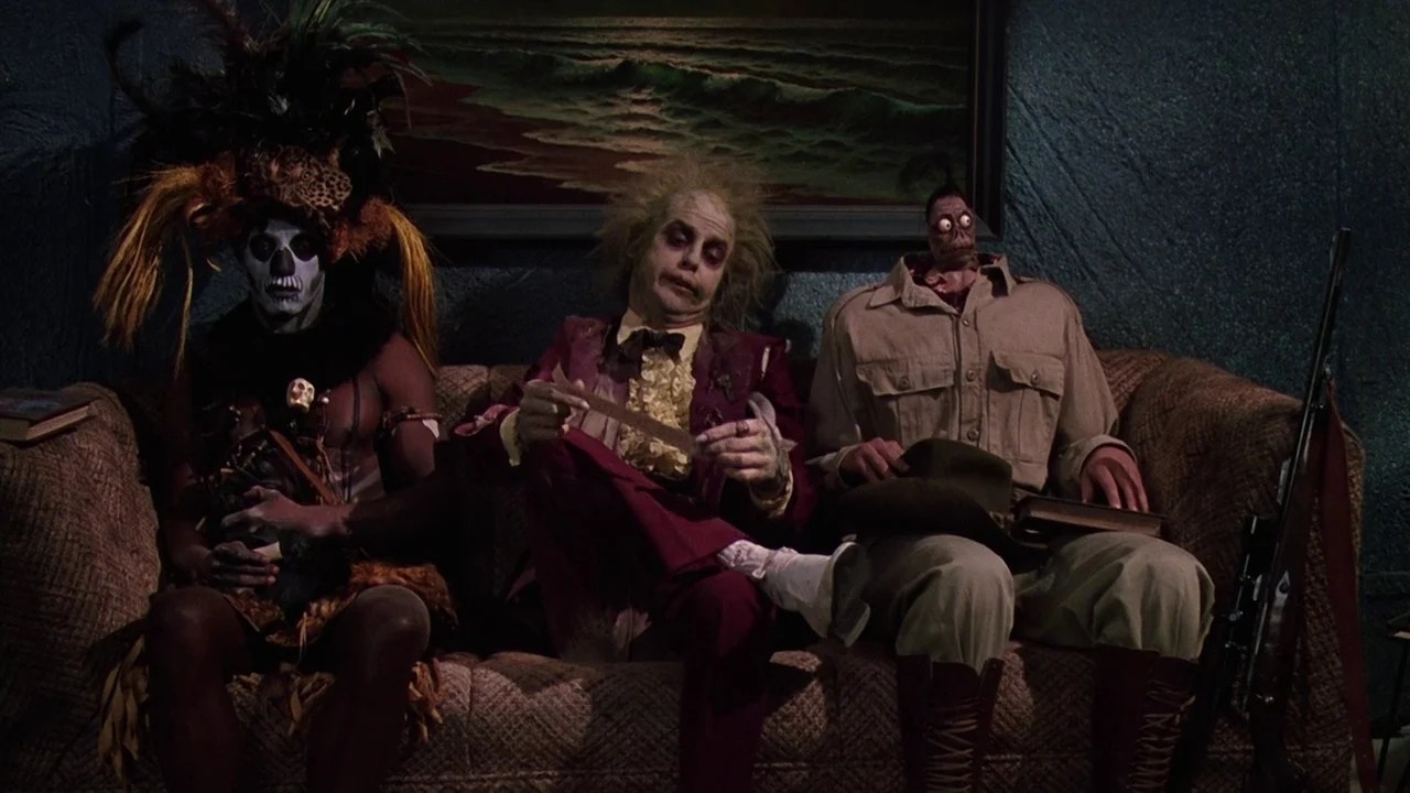 Michael Keaton as Betelgeuse, with wild hair and a burgundy suit, sitting on a sofa between a witch doctor and a man with a shrunken head in the afterlife waiting room in Beetlejuice.