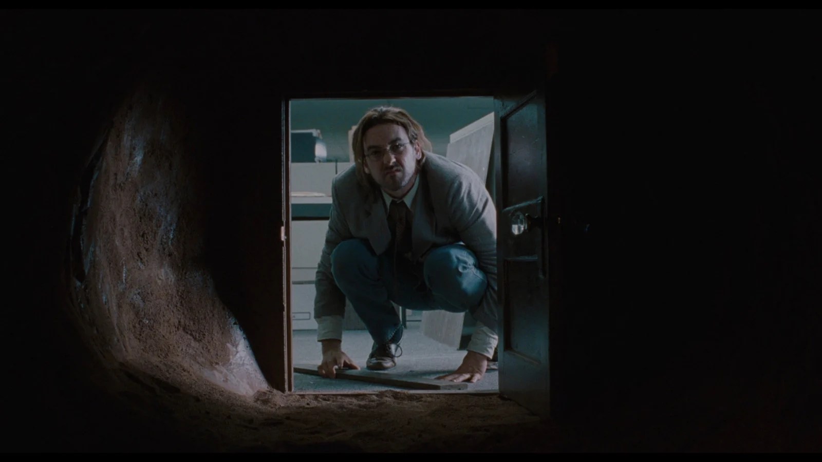 John Cusack crouching in an office doorway, peering into the dark, earthy portal in Being John Malkovich.