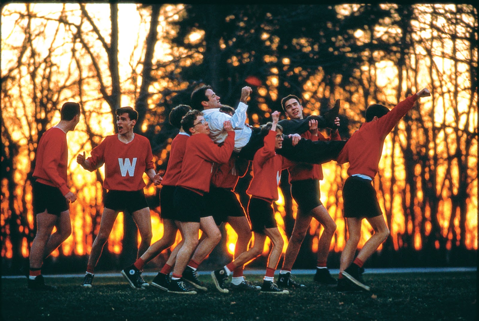 Students in red Welton sweaters lifting Robin Williams into the air in front of a glowing orange sunset through the trees in Dead Poets Society.