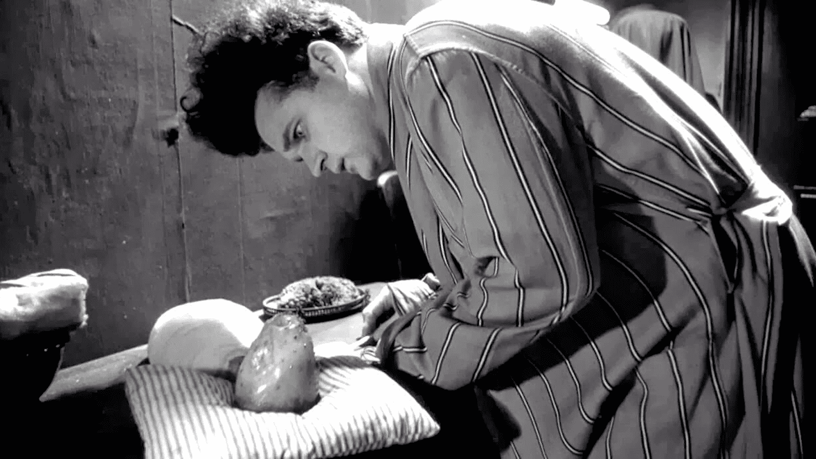 Jack Nance in a striped robe leaning over the strange swaddled baby on a pillow in a black-and-white scene from Eraserhead.