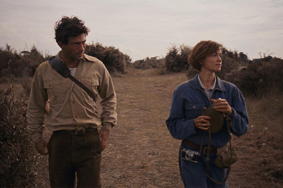 Robert Capa and Gerda Taro walk along a dry dirt path through scrubland, she in blue overalls with a camera at her waist, from the film "The Girl with the Leica".