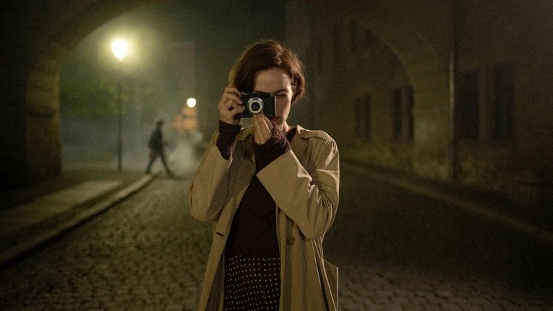 Gerda Taro, in a trench coat, raises her Leica camera to her eye on a foggy cobblestone street at night in the film The Girl with the Leica.