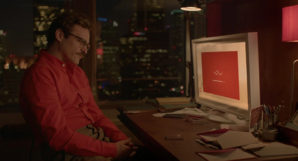 Joaquin Phoenix in a red shirt sitting at his desk at night, facing a computer screen in Her (2013).