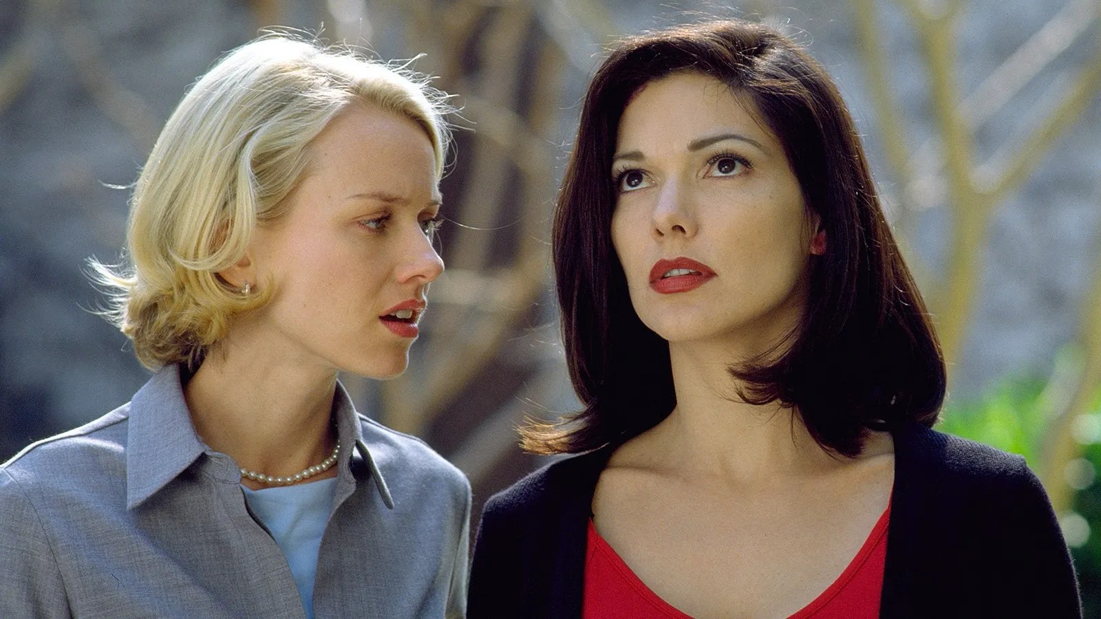 Naomi Watts, blonde in a grey jacket, talking to Laura Harring, dark-haired in red, outdoors in Mulholland Drive.
