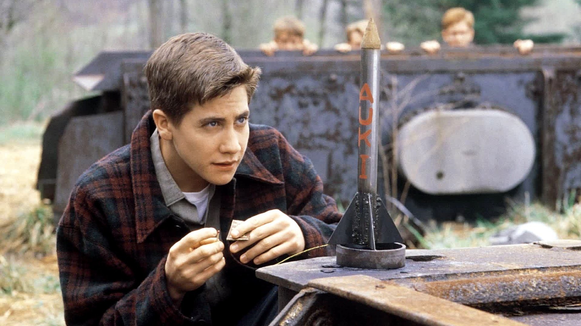 Jake Gyllenhaal in a plaid jacket lighting the fuse of a small homemade rocket while his friends watch from behind a rusty metal wall in October Sky.