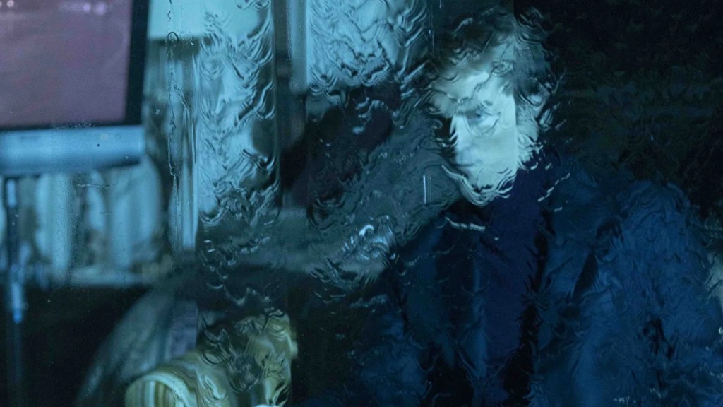 A man in a dark sweater sits beside a glowing monitor, seen through a rain-streaked window in cold blue light, from the film "Primetime".