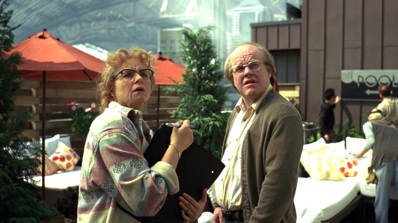 Samantha Morton as an older Hazel, holding a clipboard, beside Philip Seymour Hoffman in glasses and a cardigan, both looking up on a rooftop set in Synecdoche, New York.