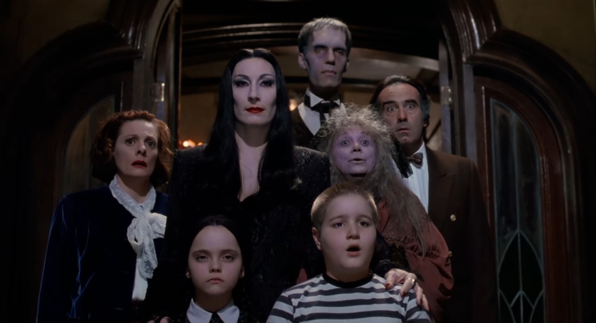 Anjelica Huston as Morticia standing behind Christina Ricci as Wednesday and Jimmy Workman as Pugsley, with Lurch, Granny and others crowded in a dark doorway, in The Addams Family (1991).