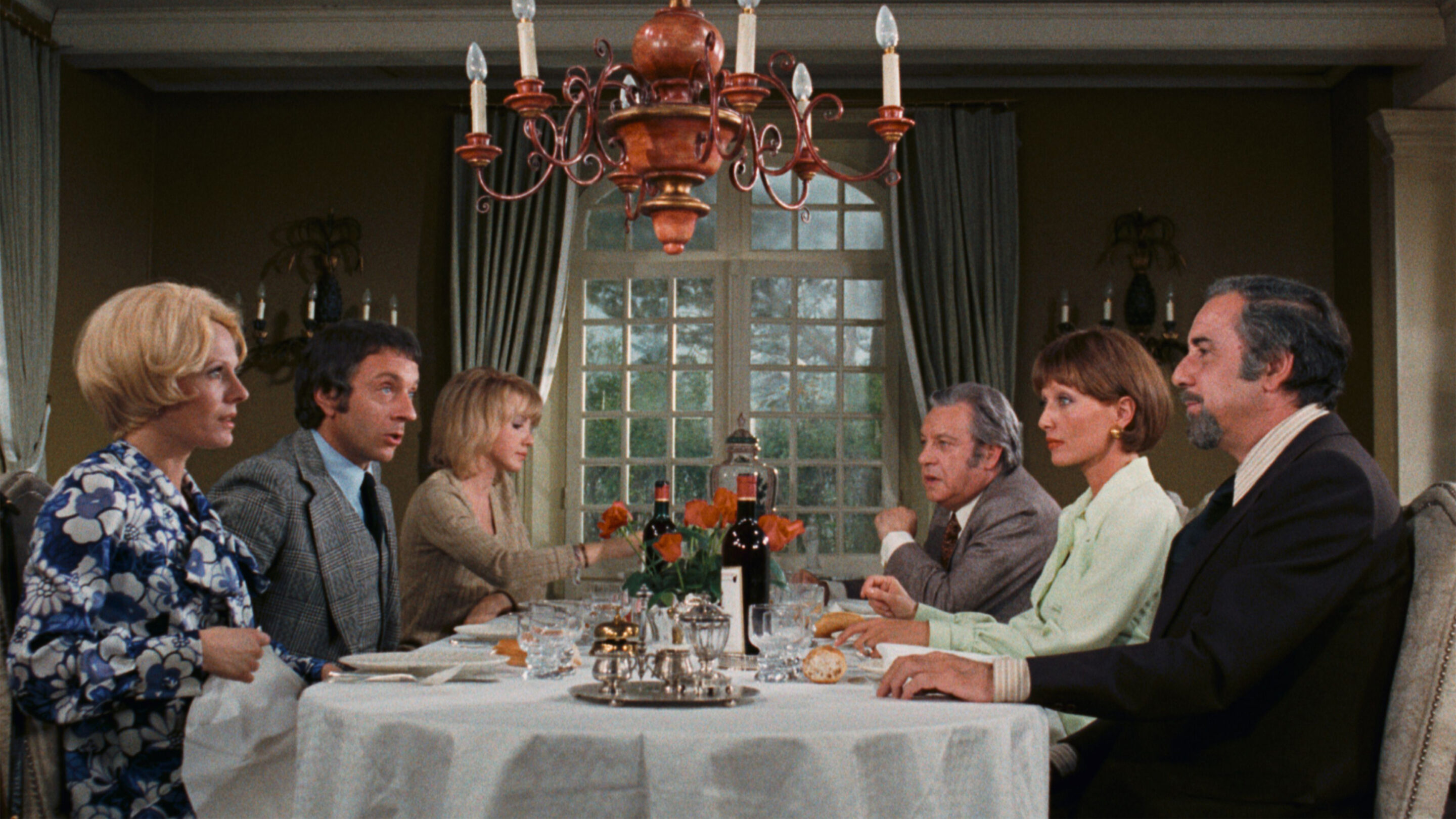 Six well-dressed friends seated around a dinner table under a chandelier in The Discreet Charm of the Bourgeoisie