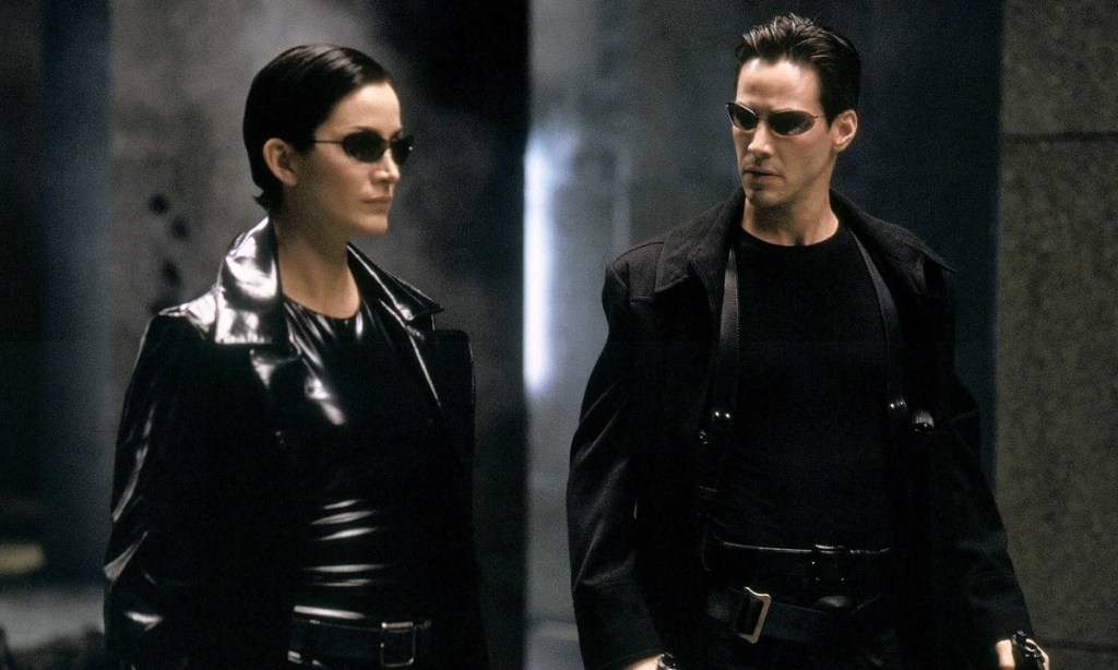 Carrie-Anne Moss and Keanu Reeves in black coats and sunglasses walking in the Matrix.