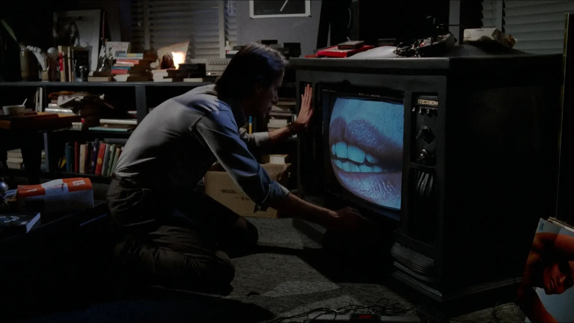James Woods kneeling in a dark room with his hand on an old TV set showing a giant pair of lips in Videodrome.